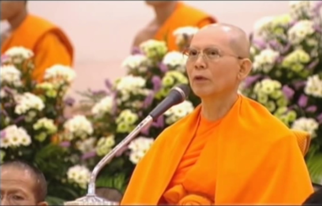 Luang Por Dhammajayo addressing an audience with flowers in the background